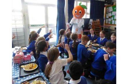 39 Mr Nourish visit. Person with orange fruit as head presenting to group of children.