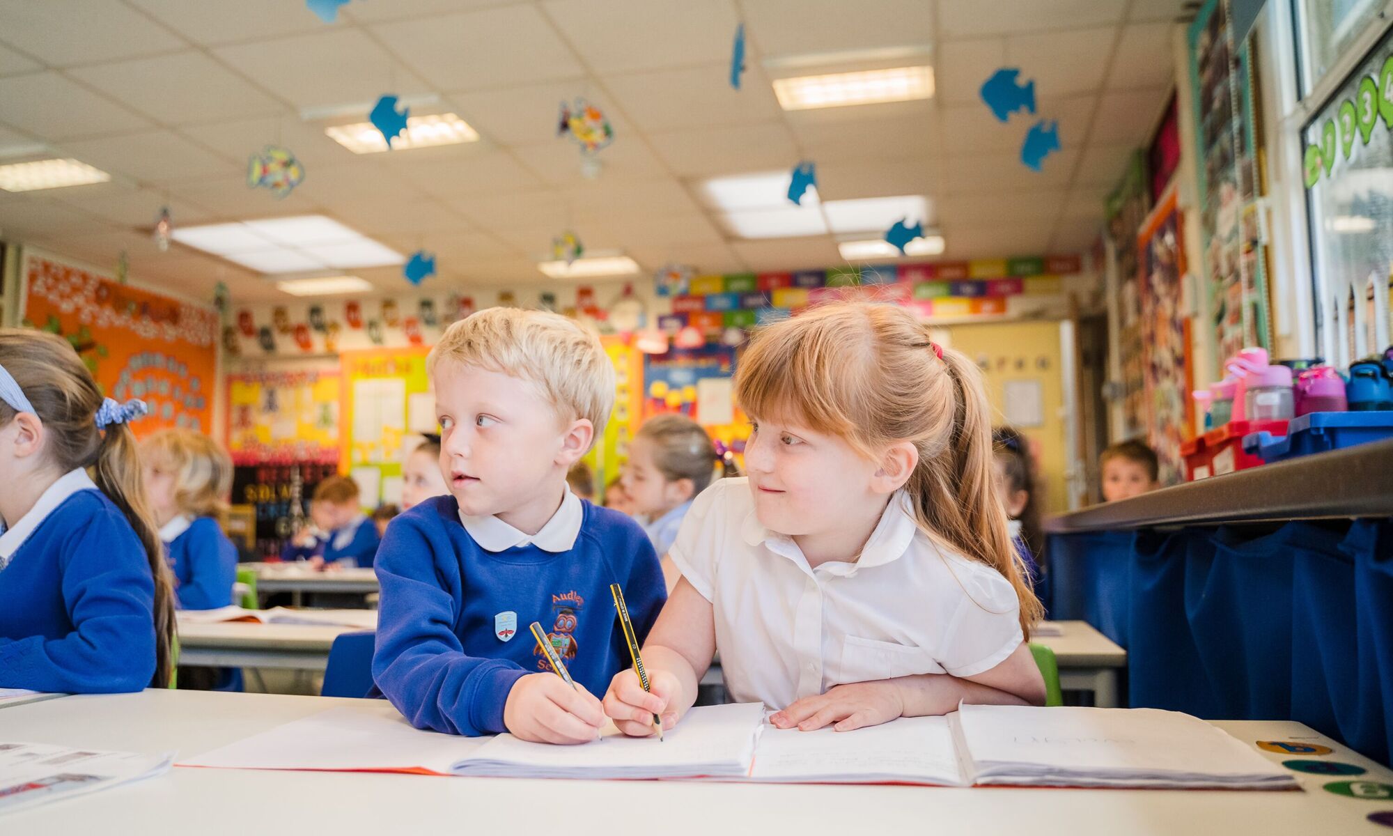 Children in a classroom. The focal point is 2 at the front working in a book